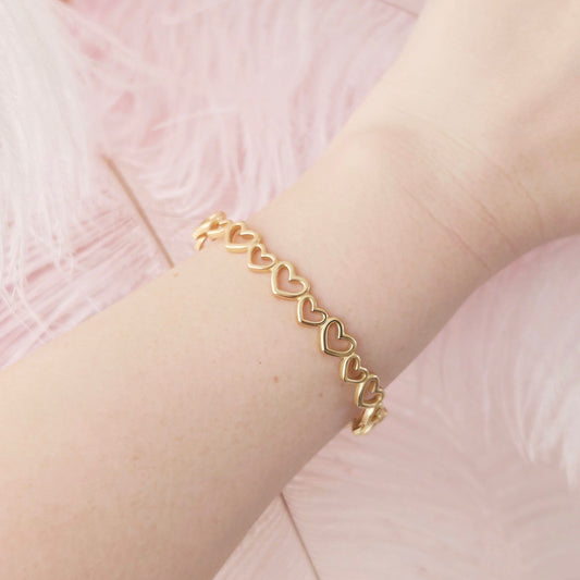 Gold bracelet with heart links on a wrist against a pink background