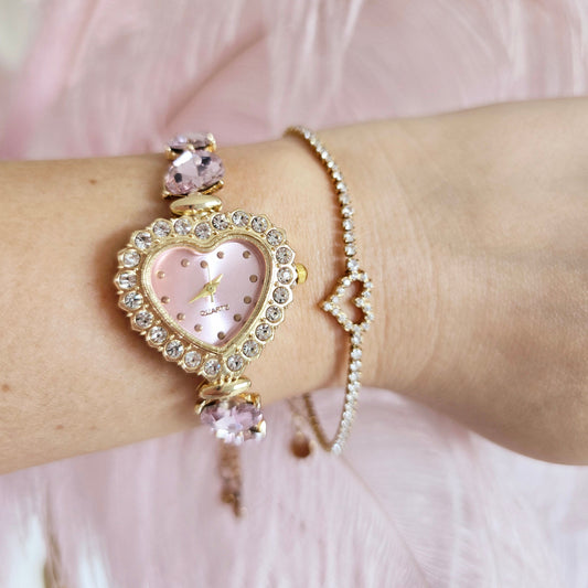 Close-up of a wrist wearing a decorative bracelet with a heart-shaped watch face against a soft pink background.