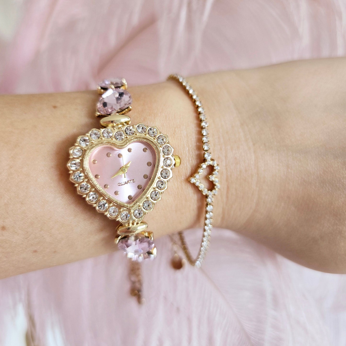 Close-up of a wrist wearing a decorative bracelet with a heart-shaped watch face against a soft pink background.